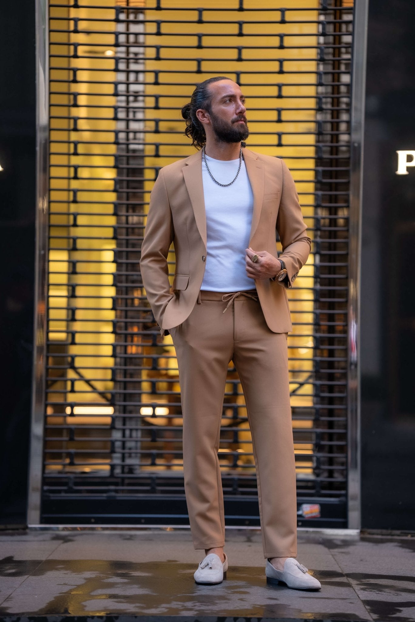 A stylish man wearing a Camel Mono Collar Slim-Fit Suit stands confidently in front of a modern storefront. The suit is paired with a simple white T-shirt, creating a relaxed yet refined look. He accessorizes with a chain necklace, a ring, and a watch, while white tassel loafers complete the ensemble. The man has a neatly groomed beard and his hair pulled back, exuding a contemporary and sophisticated vibe, perfectly balancing casual and formal elements in his outfit.