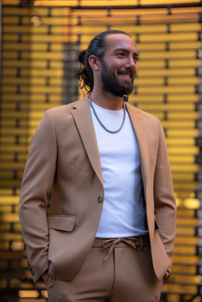 A stylish man wearing a Camel Mono Collar Slim-Fit Suit stands confidently in front of a modern storefront. The suit is paired with a simple white T-shirt, creating a relaxed yet refined look. He accessorizes with a chain necklace, a ring, and a watch, while white tassel loafers complete the ensemble. The man has a neatly groomed beard and his hair pulled back, exuding a contemporary and sophisticated vibe, perfectly balancing casual and formal elements in his outfit.