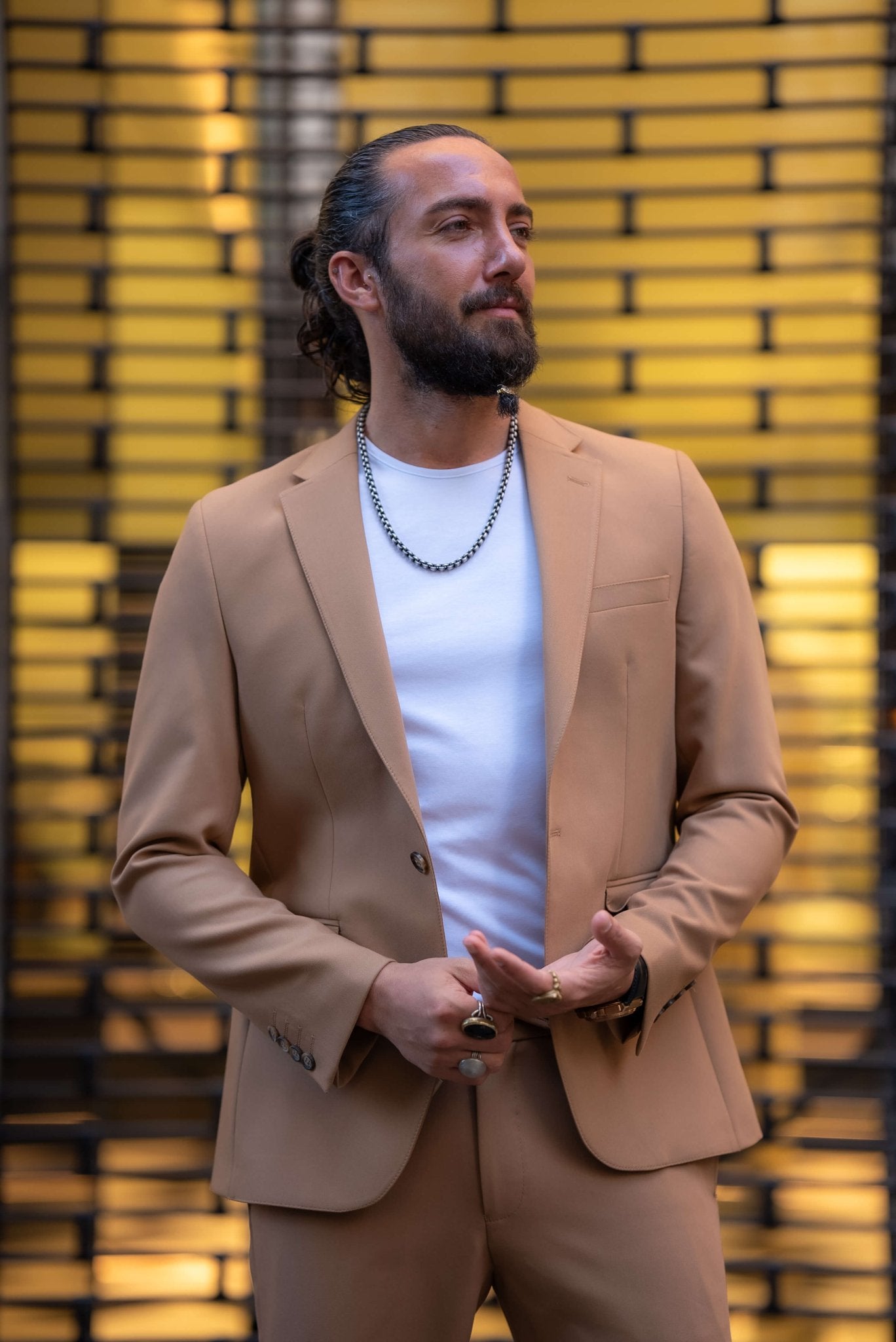 A stylish man wearing a Camel Mono Collar Slim-Fit Suit stands confidently in front of a modern storefront. The suit is paired with a simple white T-shirt, creating a relaxed yet refined look. He accessorizes with a chain necklace, a ring, and a watch, while white tassel loafers complete the ensemble. The man has a neatly groomed beard and his hair pulled back, exuding a contemporary and sophisticated vibe, perfectly balancing casual and formal elements in his outfit.