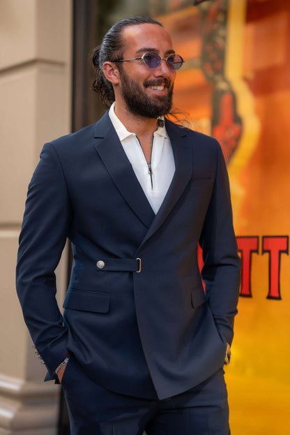 A stylish man wearing a Navy Blue Single Lapel Button Suit leans casually against a building in an urban setting. The suit features a unique design with a single lapel and silver button closure, paired with a white shirt and white sneakers, blending modern sophistication with a casual edge. The man accessorizes with multiple rings, a watch, and round sunglasses, completing his contemporary and chic look. His neatly groomed beard and relaxed posture enhance the effortless elegance of his outfit.