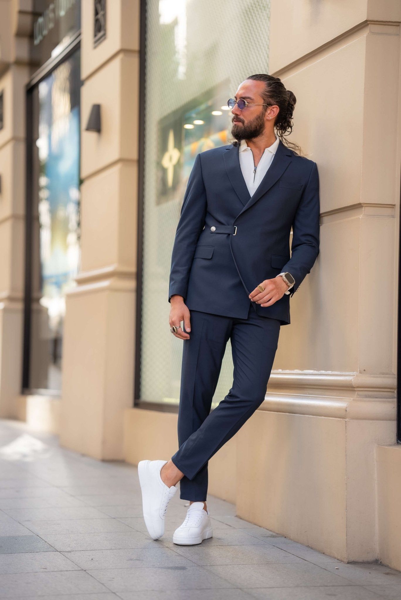 A stylish man wearing a Navy Blue Single Lapel Button Suit leans casually against a building in an urban setting. The suit features a unique design with a single lapel and silver button closure, paired with a white shirt and white sneakers, blending modern sophistication with a casual edge. The man accessorizes with multiple rings, a watch, and round sunglasses, completing his contemporary and chic look. His neatly groomed beard and relaxed posture enhance the effortless elegance of his outfit.