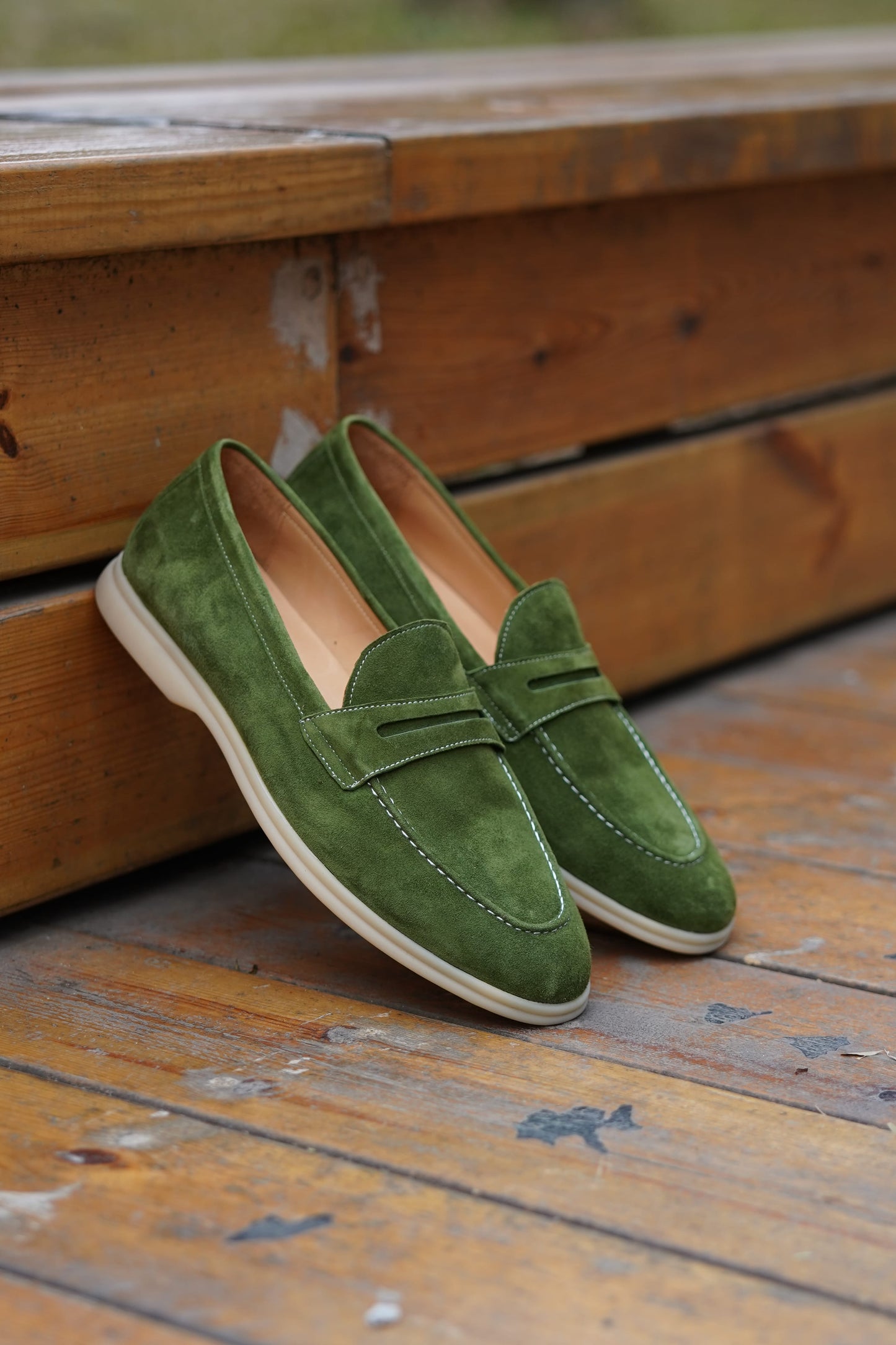 Hand holding olive green suede penny loafers outdoors, three-quarter view