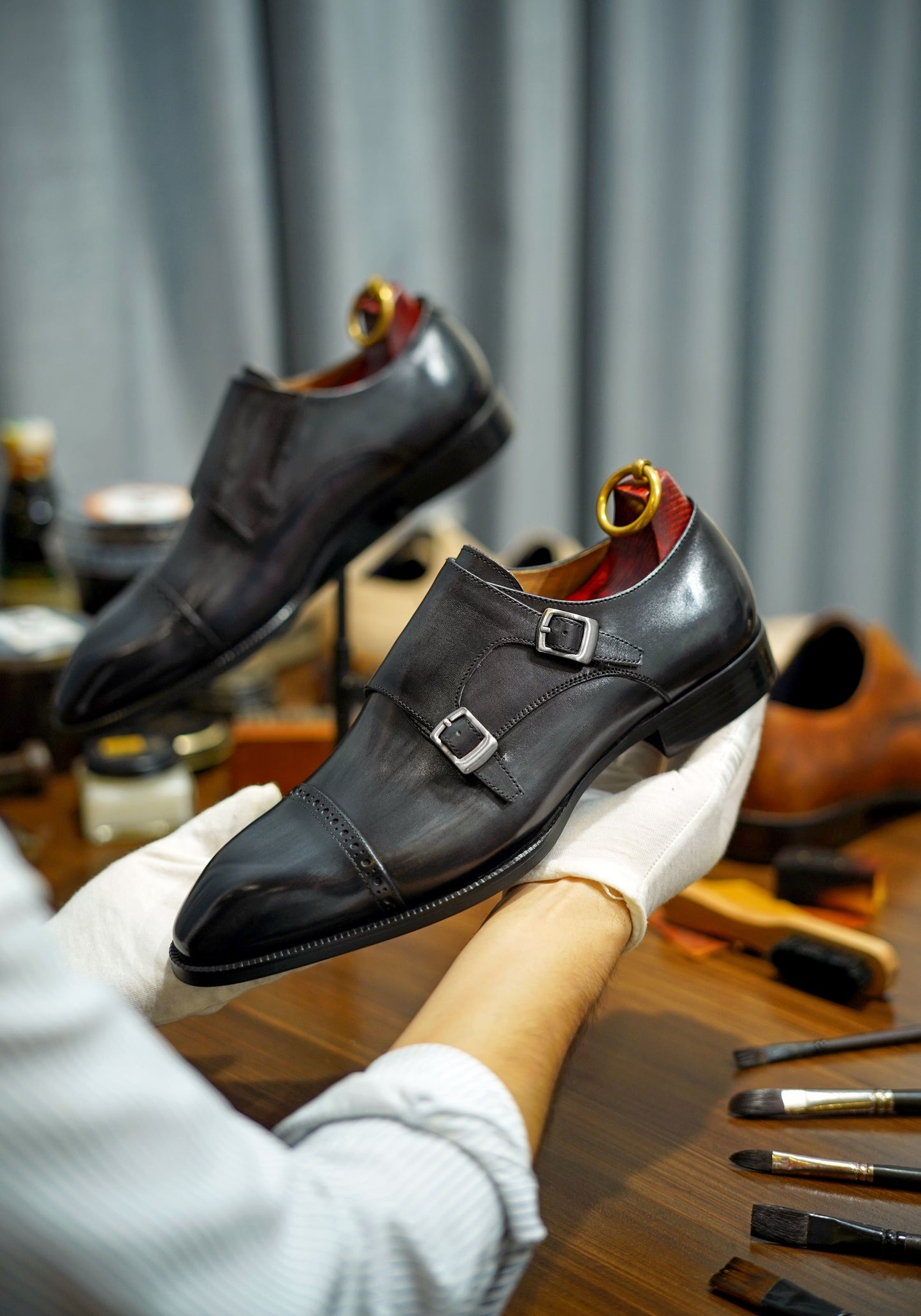 Polished Black Double Monk Strap Leather Shoes – pair held by gloved hands, emphasizing the sophisticated design and craftsmanship with shoe polish tools in the background.