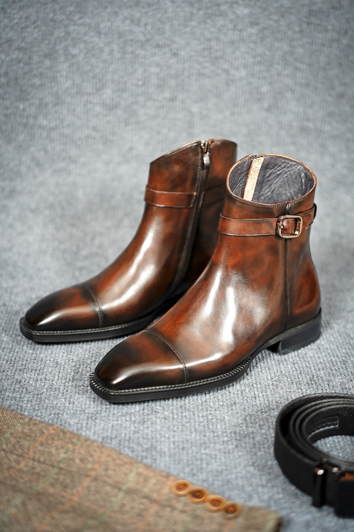 Premium Brown Leather Ankle Boots for Urban Adventures – a pair of brown leather boots on a gray textured background, with a belt and checkered fabric.