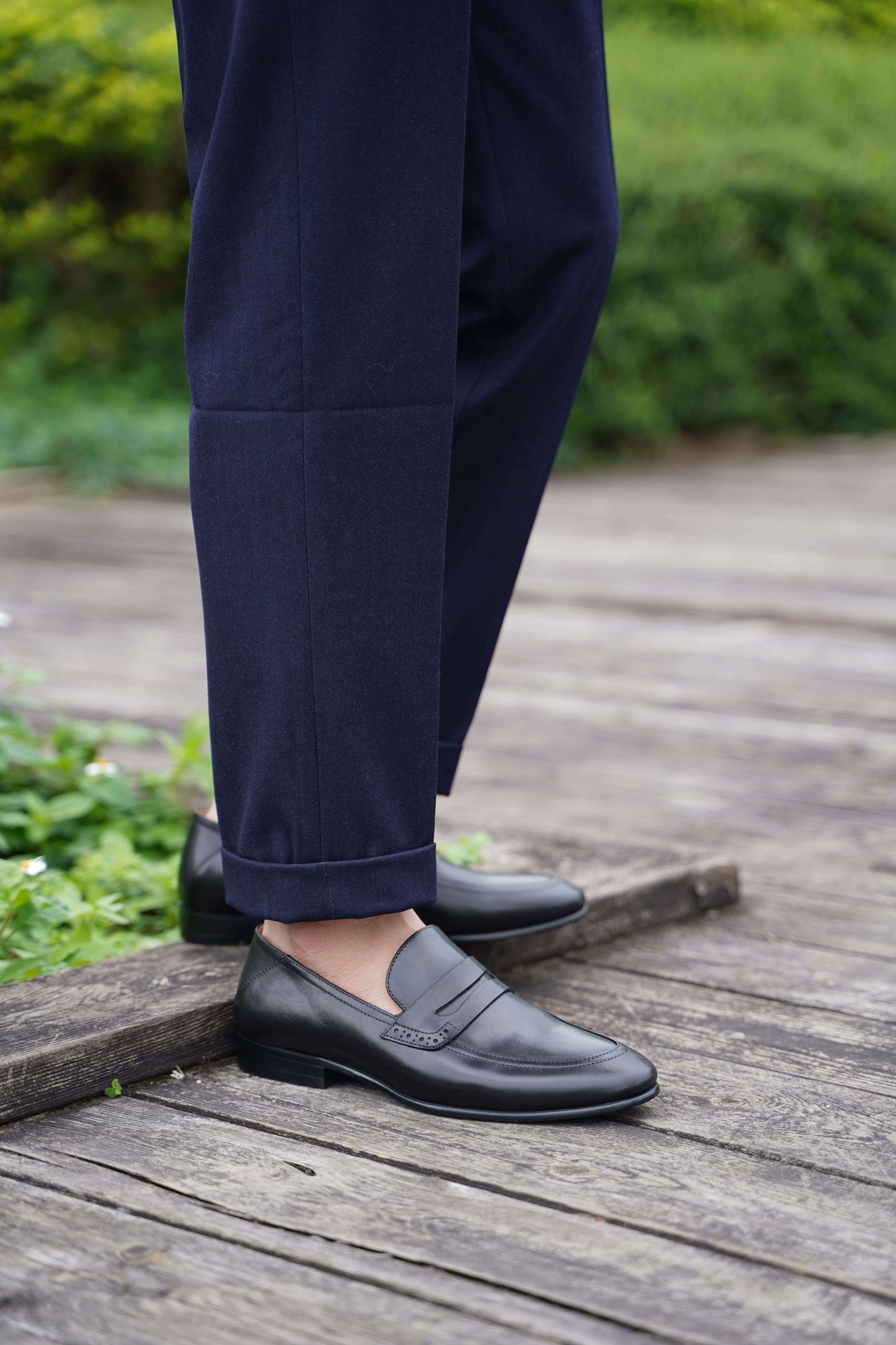 Refined Black Leather Penny Loafers – worn with cuffed navy trousers on a wooden deck, showcasing timeless sophistication and a polished finish.