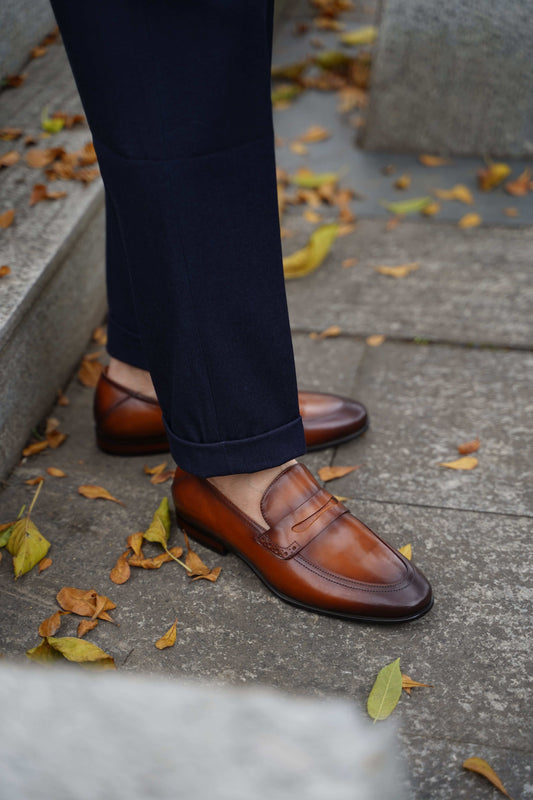 Brown Leather Penny Loafers – worn by a man in cuffed navy trousers, highlighting sleek silhouette and a polished finish on a paved surface.