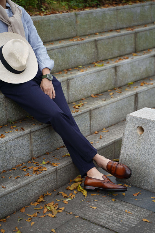Refined Brown Leather Penny Loafers – worn by a man in navy trousers, sitting on steps with a straw hat, showcasing timeless elegance.