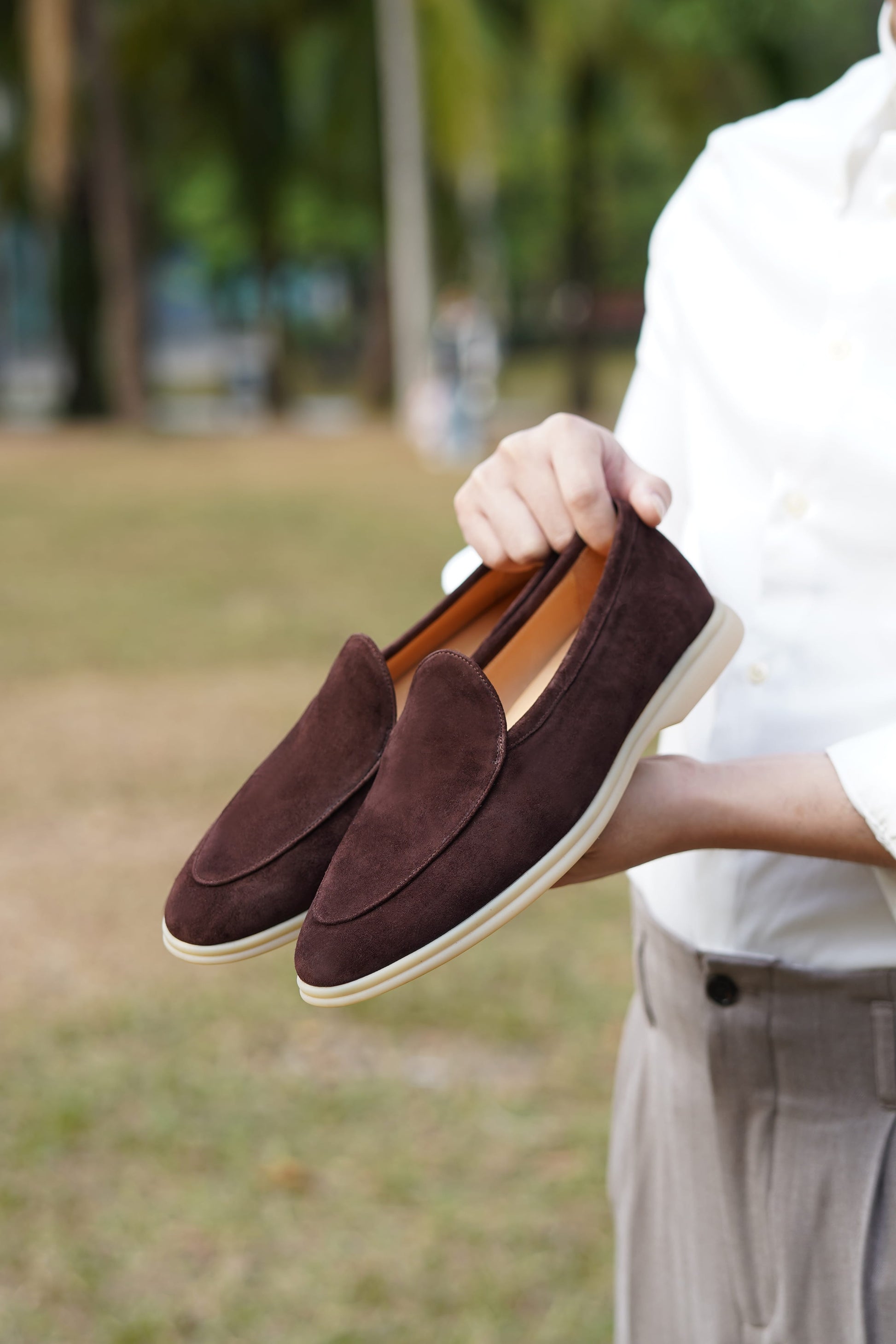 Hand holding Siena coffee-brown suede Venetian loafers outdoors, three-quarter view