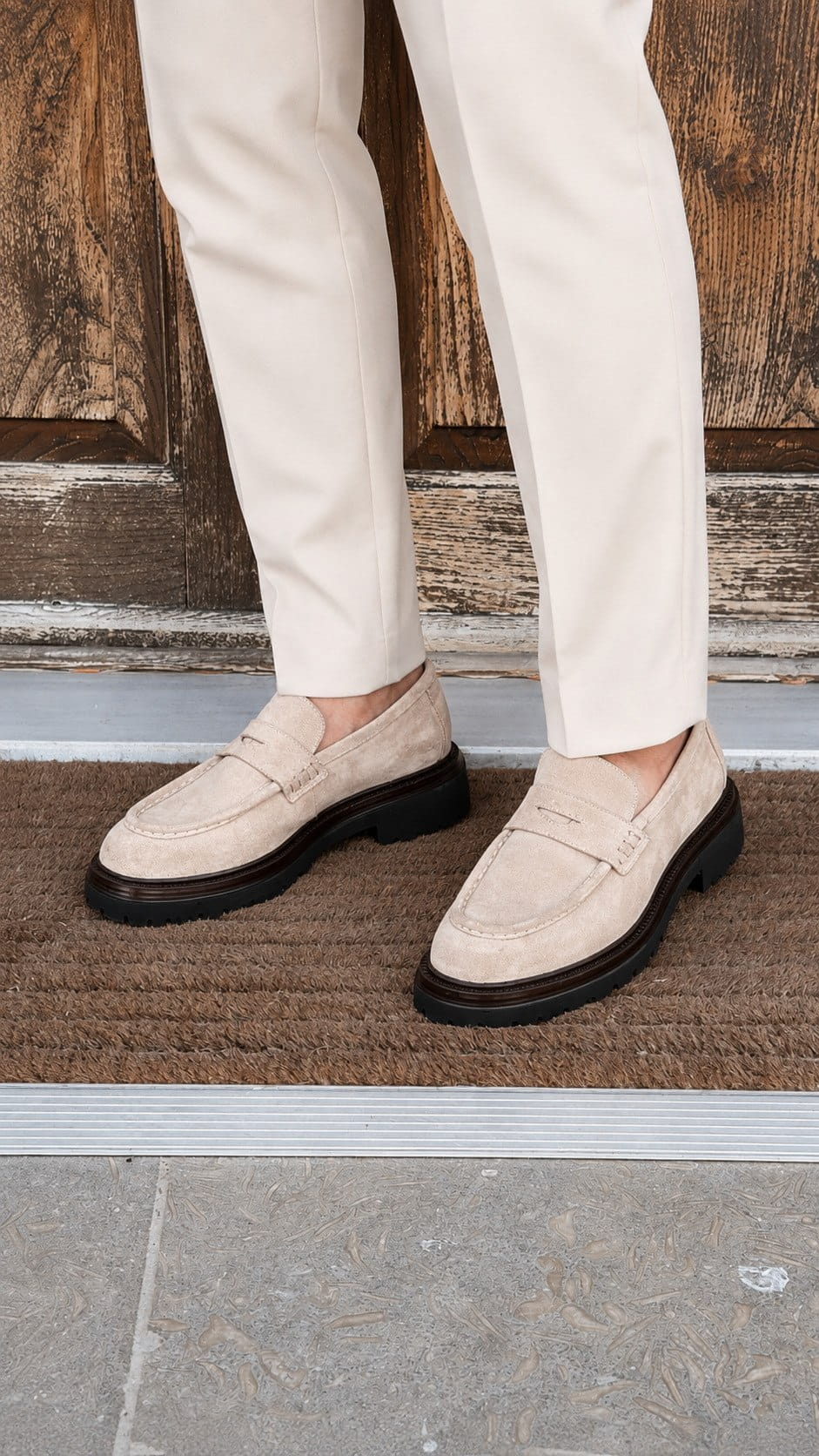 A person wearing off-white trousers and light beige suede penny loafers with black soles, standing on a brown doormat in front of a rustic wooden door.