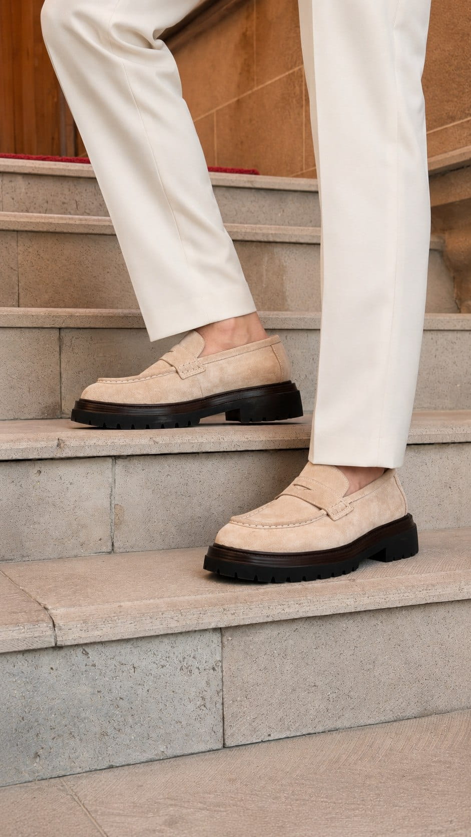 A person wearing off-white trousers and beige suede penny loafers with chunky black soles, standing on stone stairs.