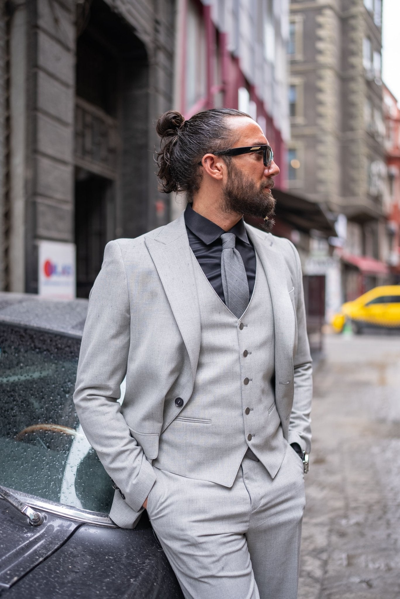 A stylish man wearing a Signature Gray Slim Fit Suit stands confidently by a vintage car on a city street. The suit is paired with a black dress shirt, a gray tie, and black loafers, exuding a modern and sophisticated look. The man has a neatly groomed beard and wears sunglasses, adding to his dapper appearance.