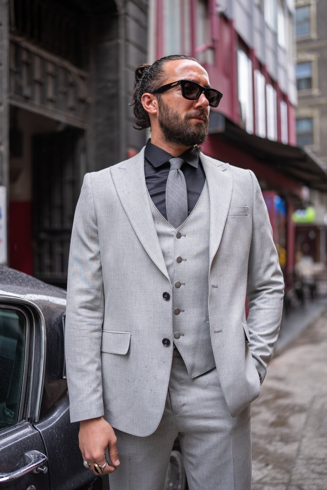 A stylish man wearing a Signature Gray Slim Fit Suit stands confidently by a vintage car on a city street. The suit is paired with a black dress shirt, a gray tie, and black loafers, exuding a modern and sophisticated look. The man has a neatly groomed beard and wears sunglasses, adding to his dapper appearance.