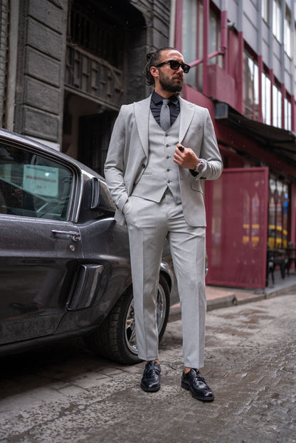 A stylish man wearing a Signature Gray Slim Fit Suit stands confidently by a vintage car on a city street. The suit is paired with a black dress shirt, a gray tie, and black loafers, exuding a modern and sophisticated look. The man has a neatly groomed beard and wears sunglasses, adding to his dapper appearance.