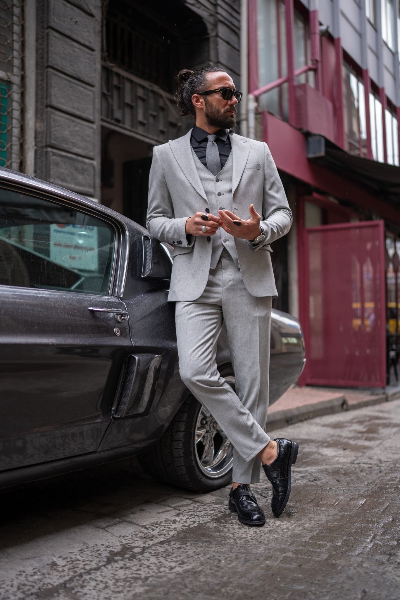 A stylish man wearing a Signature Gray Slim Fit Suit stands confidently by a vintage car on a city street. The suit is paired with a black dress shirt, a gray tie, and black loafers, exuding a modern and sophisticated look. The man has a neatly groomed beard and wears sunglasses, adding to his dapper appearance.