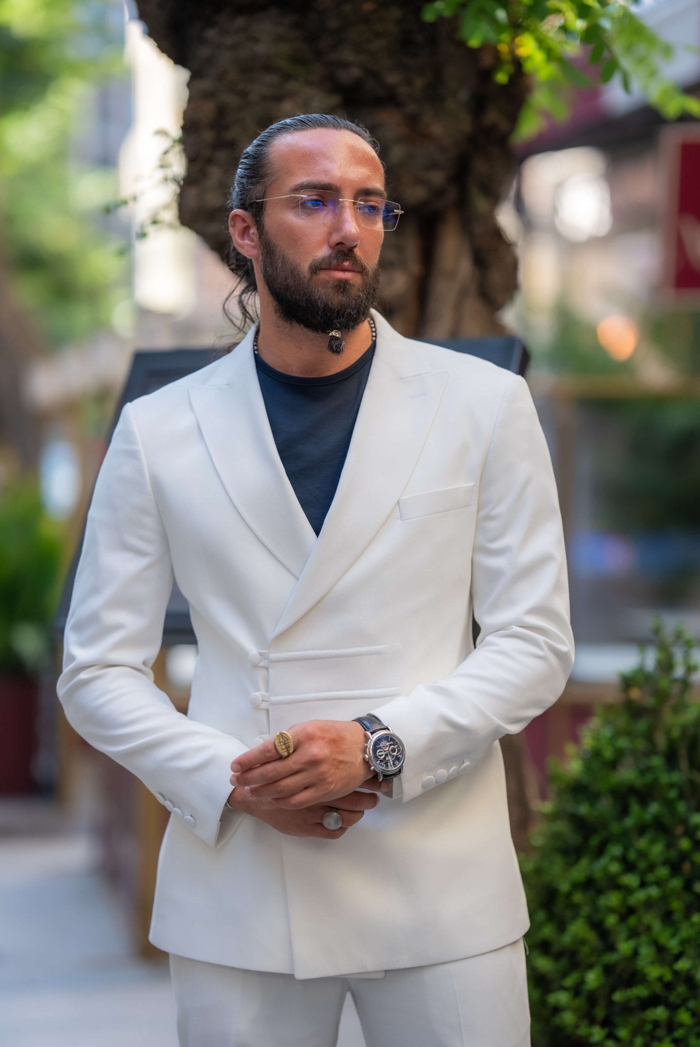 A stylish man wearing an Ecru White Mono Collar Suit leans casually against a glass partition in an outdoor setting. The suit is paired with a dark shirt and sleek navy slip-on sneakers, creating a clean and modern look. The man accessorizes with glasses, a watch, and a ring, adding subtle elegance to his outfit. His neatly groomed beard and relaxed posture enhance the sophisticated yet laid-back vibe of the ensemble, making a strong statement in contemporary fashion.