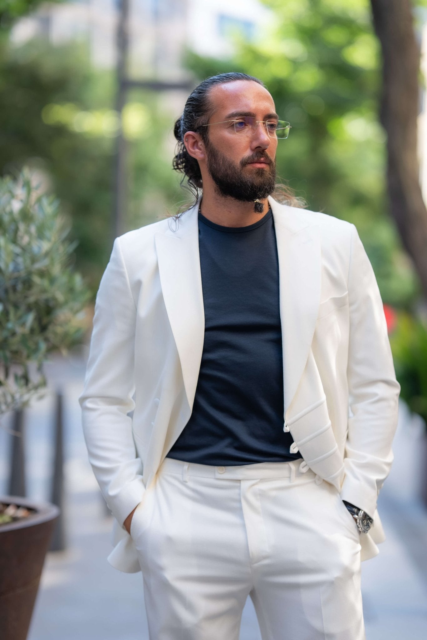 A stylish man wearing an Ecru White Mono Collar Suit leans casually against a glass partition in an outdoor setting. The suit is paired with a dark shirt and sleek navy slip-on sneakers, creating a clean and modern look. The man accessorizes with glasses, a watch, and a ring, adding subtle elegance to his outfit. His neatly groomed beard and relaxed posture enhance the sophisticated yet laid-back vibe of the ensemble, making a strong statement in contemporary fashion.