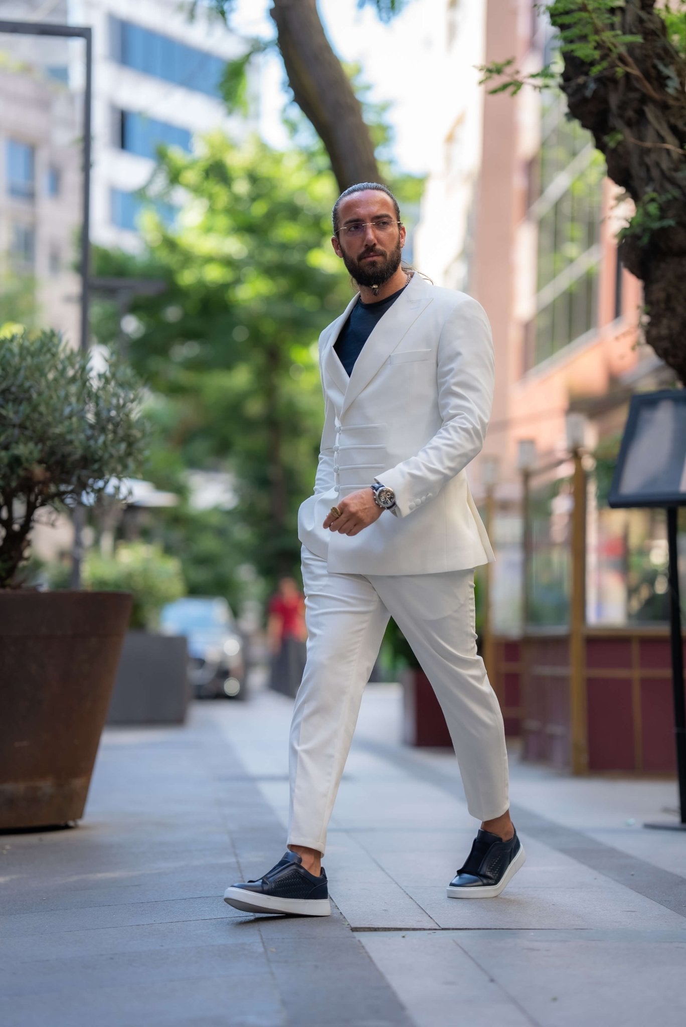 A stylish man wearing an Ecru White Mono Collar Suit leans casually against a glass partition in an outdoor setting. The suit is paired with a dark shirt and sleek navy slip-on sneakers, creating a clean and modern look. The man accessorizes with glasses, a watch, and a ring, adding subtle elegance to his outfit. His neatly groomed beard and relaxed posture enhance the sophisticated yet laid-back vibe of the ensemble, making a strong statement in contemporary fashion.