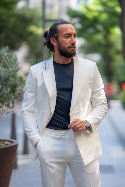 A stylish man wearing an Ecru White Mono Collar Suit leans casually against a glass partition in an outdoor setting. The suit is paired with a dark shirt and sleek navy slip-on sneakers, creating a clean and modern look. The man accessorizes with glasses, a watch, and a ring, adding subtle elegance to his outfit. His neatly groomed beard and relaxed posture enhance the sophisticated yet laid-back vibe of the ensemble, making a strong statement in contemporary fashion.