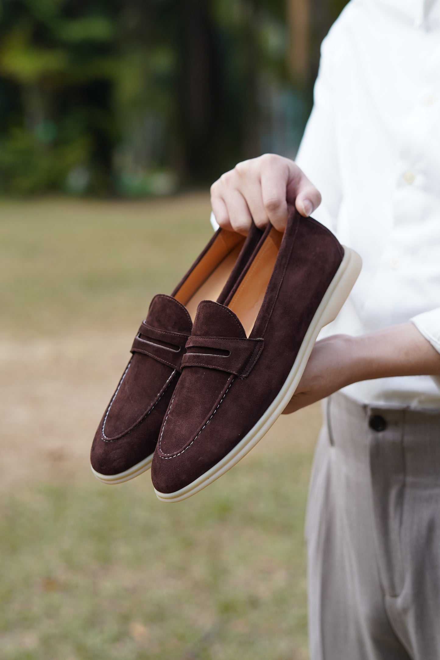 Hand holding espresso-brown suede penny loafers outdoors, three-quarter view