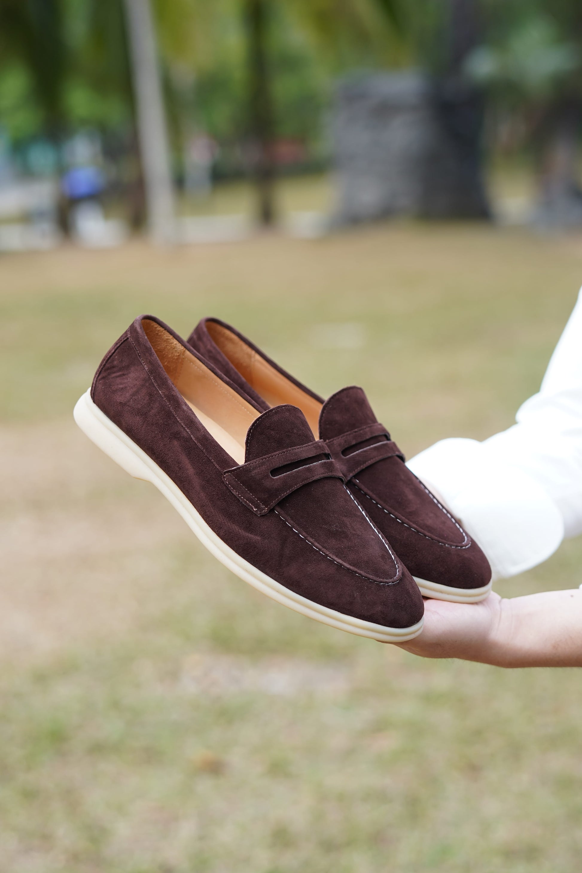 Espresso-brown suede penny loafers held against greenery, detail view