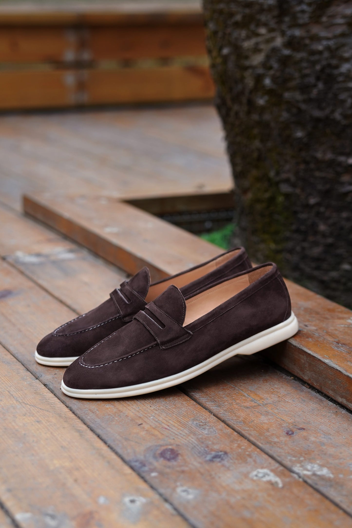 Espresso-brown suede penny loafers on a textured wooden surface, angled view