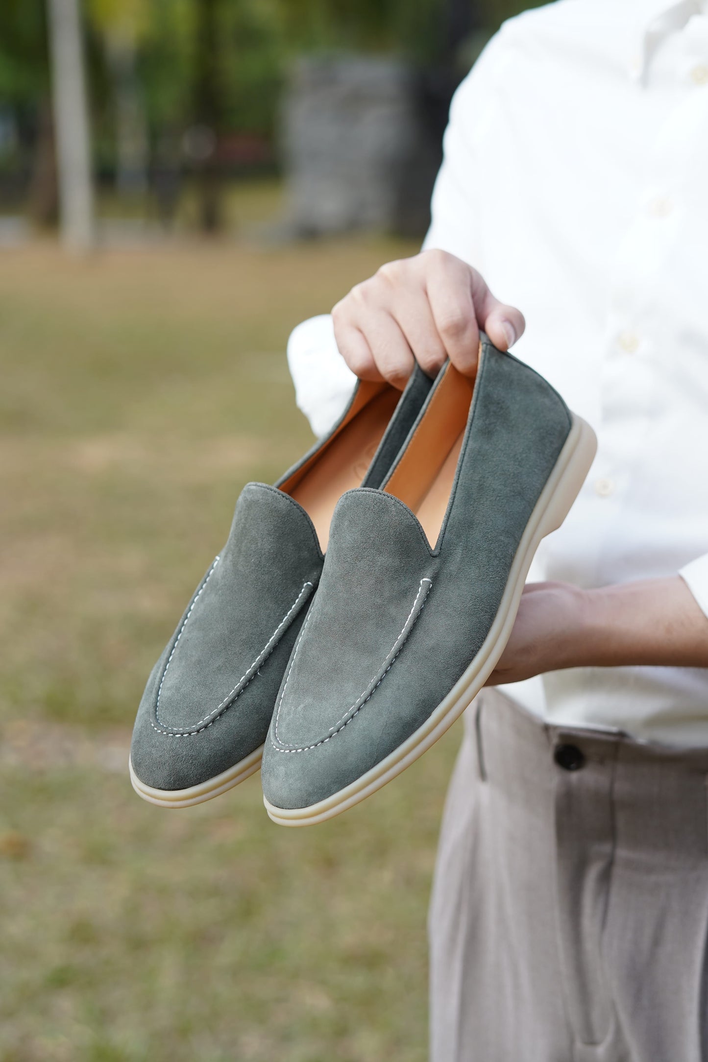 Sage gray suede Venetian loafers held outdoors against greenery, detail view