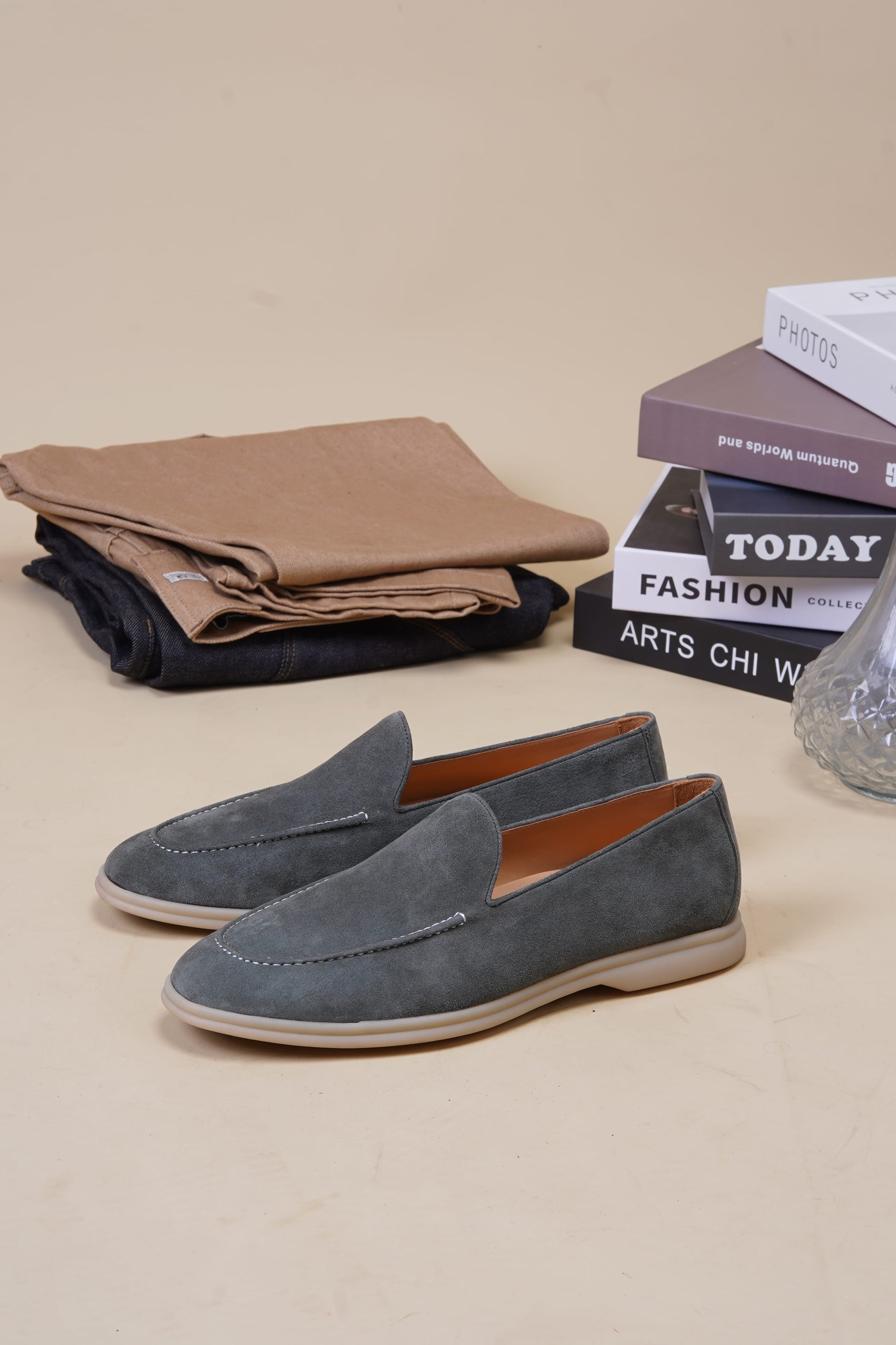 Sage gray suede Venetian loafers on beige surface with folded clothes and books, angled pair