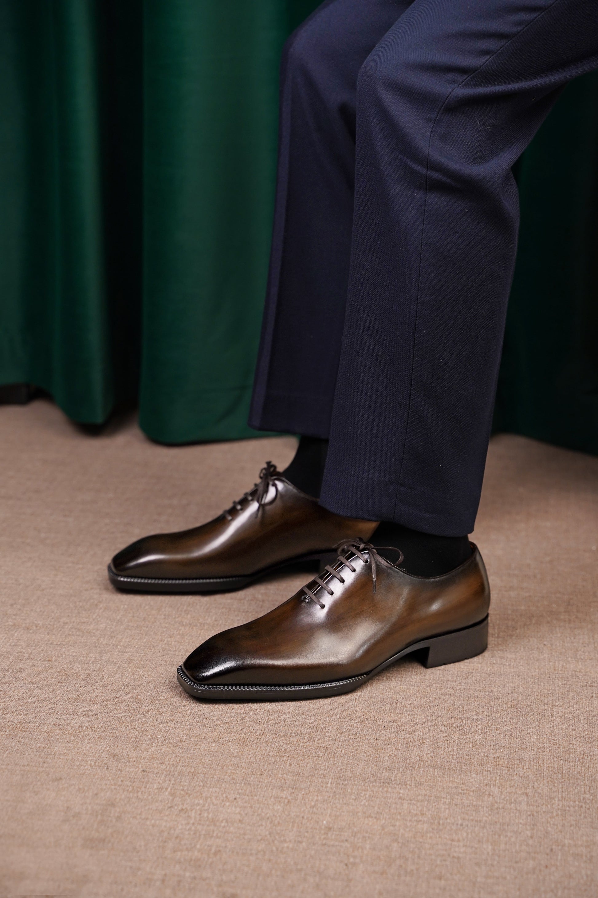 The Regent Cognac Wholecut Oxford– a view of the shoes being worn by a person in dark blue formal trousers, standing on a light-colored carpet.