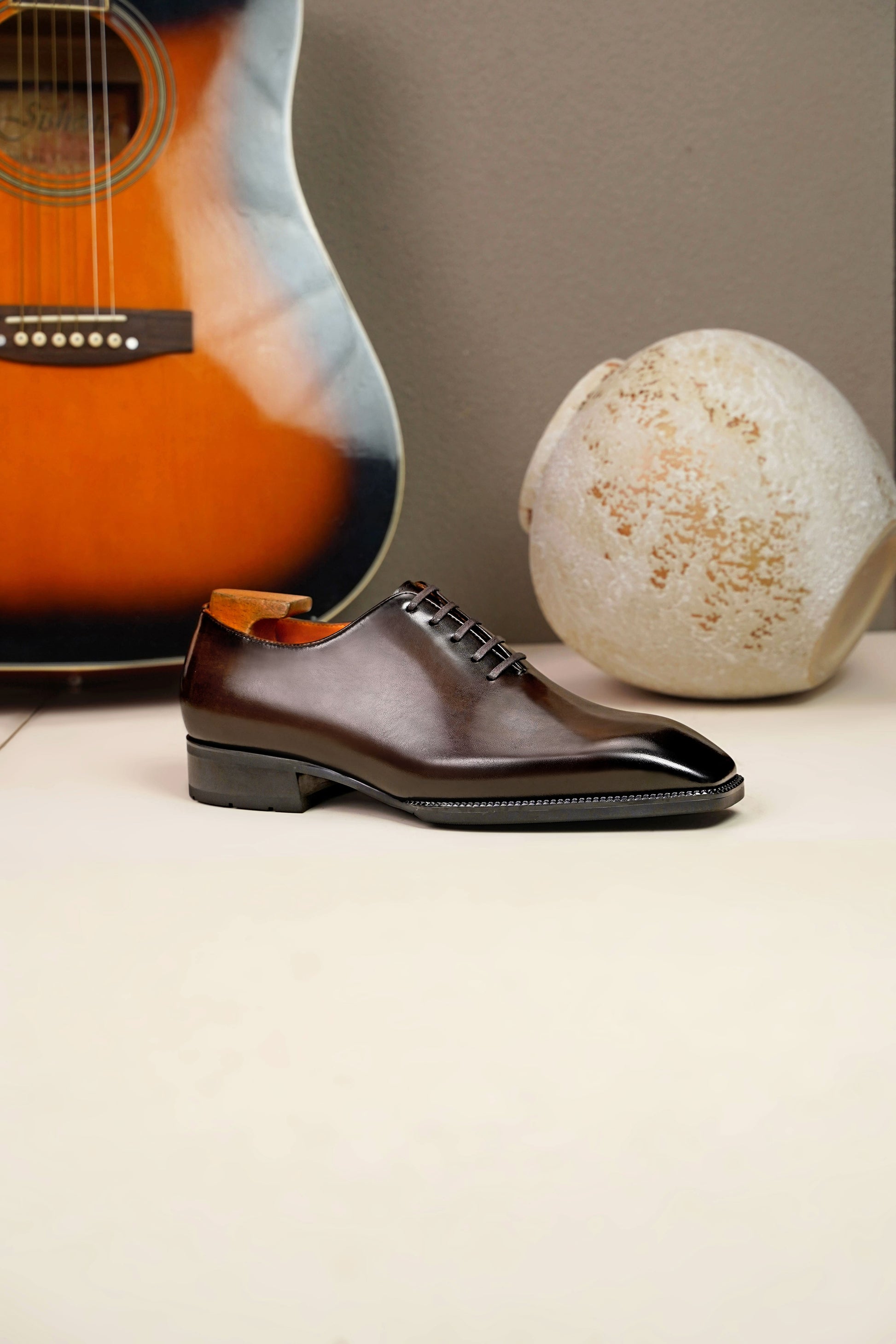 The Regent Cognac Wholecut Oxford: a side-profile view of a single shoe standing on a light surface, with an acoustic guitar and an artistic vase in the background.