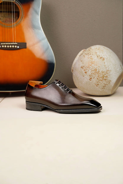 The Regent Cognac Wholecut Oxford: a side-profile view of a single shoe standing on a light surface, with an acoustic guitar and an artistic vase in the background.