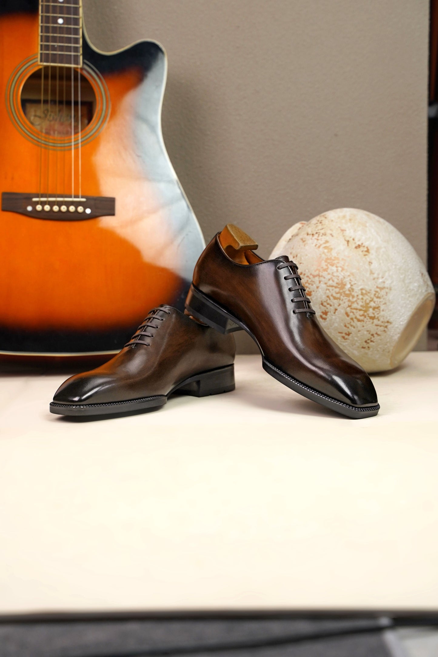 The Regent Cognac Wholecut Oxford – a dynamic arrangement of the two shoes on a light surface, with one shoe propped against a wooden vase and the other resting on a shoe brush.