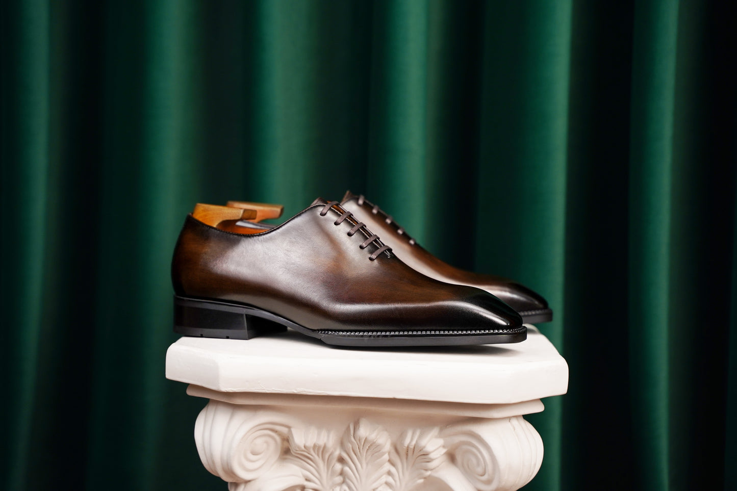 The Regent Cognac Wholecut Oxford – a side-profile shot of a pair of elegant footwear on a white classical pedestal against a dark green velvet curtain backdrop.