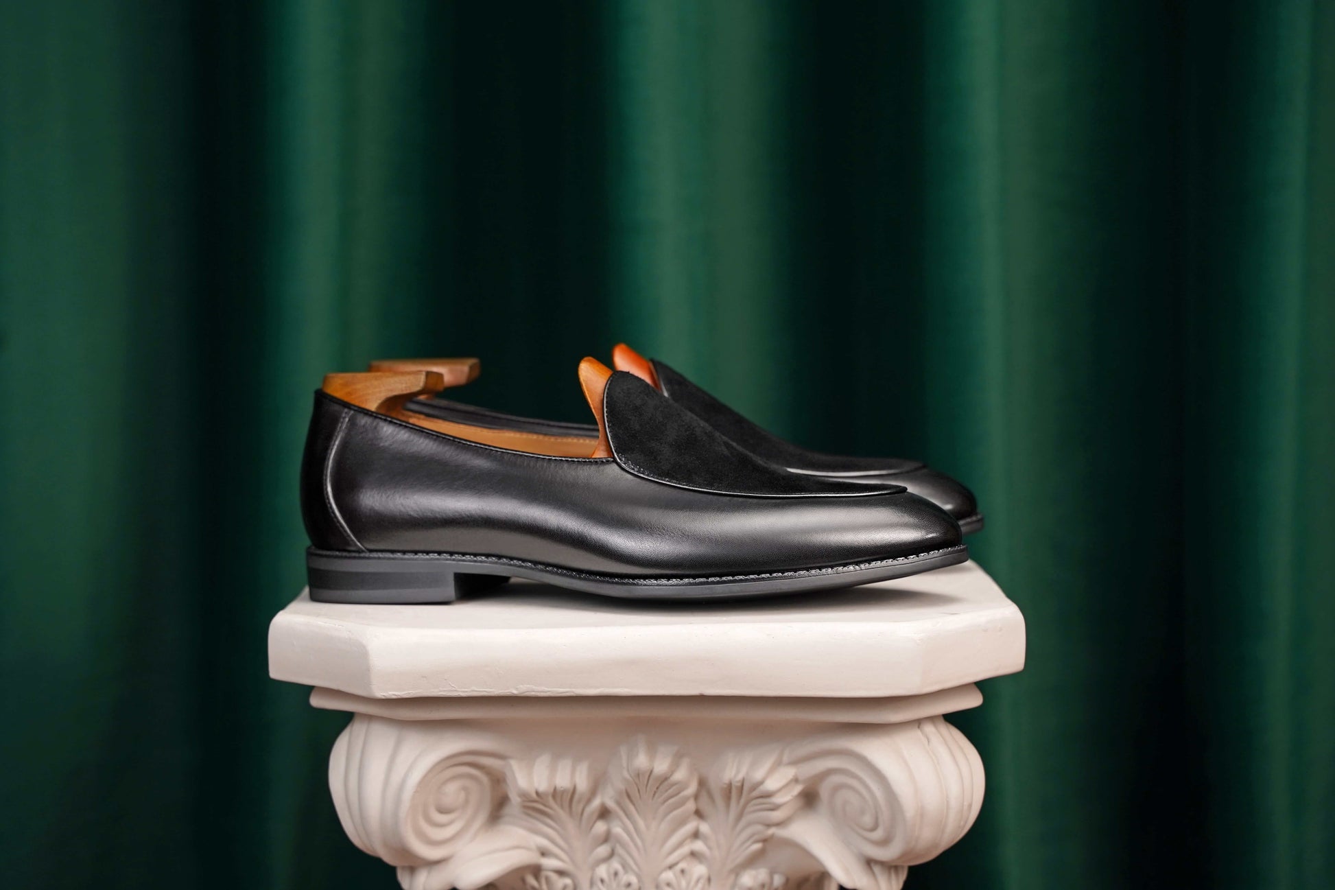 Velvet Crest Black Leather Suede Loafers – pair with shoe trees, resting on a white classical pedestal against a green curtain, showcasing the contrast in textures for refined ease.
