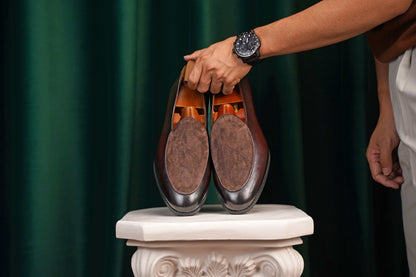 Velvet Crest Brown Leather Suede Loafers – pair held by a man's hand with a watch, showcasing the contrast between the brown leather and suede, ideal for a summer tuxedo.