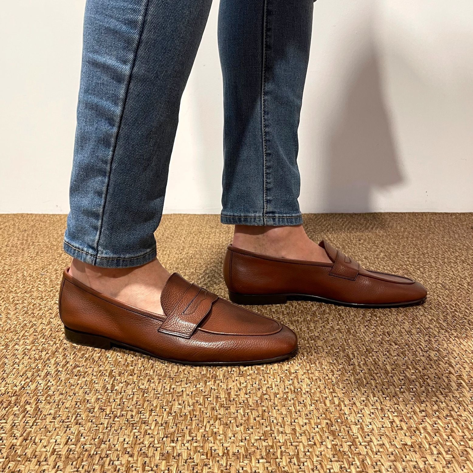 A person wearing blue jeans and brown leather penny loafers standing on a woven tan rug against a plain light wall.