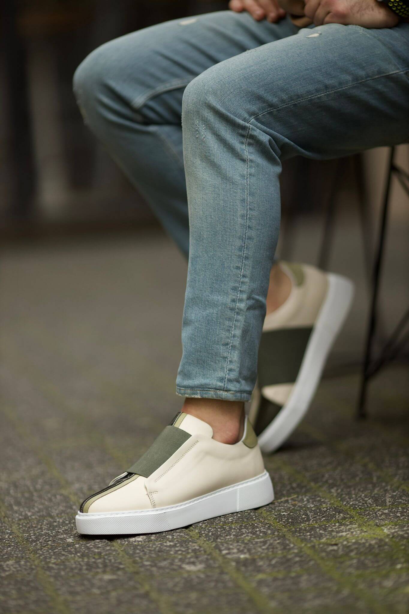 Vince Beige & Green Slip-On Sneakers - Sophisticated Comfort for Every Occasion – a man sitting in blue jeans, with one foot elevated, highlighting the two-tone leather and slip-on style.