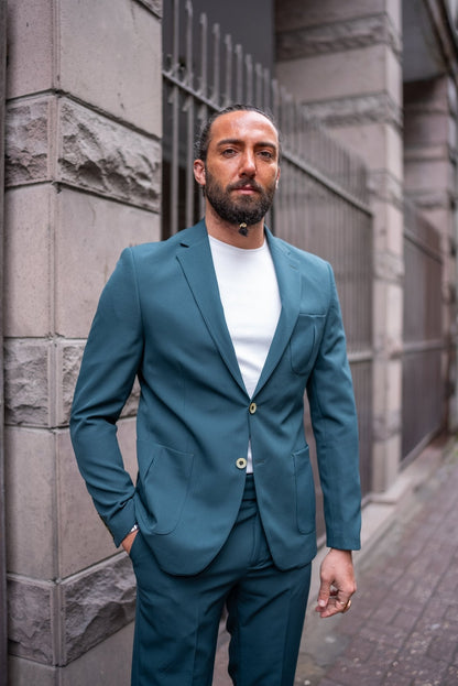 A man confidently models an Emerald Slim Fit Wedding-Casual Suit. The suit features a modern design with a sharp fit, highlighted by its vibrant emerald green color. He pairs the suit with a simple white shirt underneath, exuding a blend of elegance and casual sophistication. The man is accessorized with a large black ring, a watch, and minimal jewelry, standing in front of a stone building, capturing the essence of contemporary style.