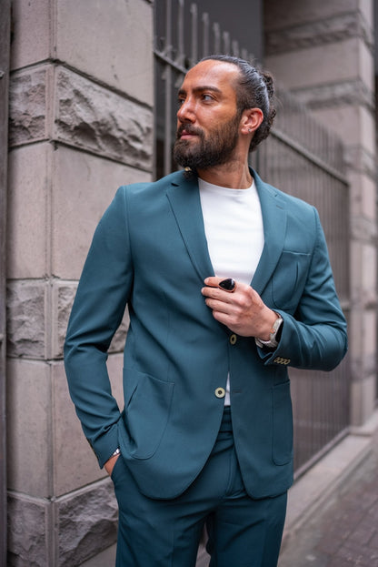 A man confidently models an Emerald Slim Fit Wedding-Casual Suit. The suit features a modern design with a sharp fit, highlighted by its vibrant emerald green color. He pairs the suit with a simple white shirt underneath, exuding a blend of elegance and casual sophistication. The man is accessorized with a large black ring, a watch, and minimal jewelry, standing in front of a stone building, capturing the essence of contemporary style.