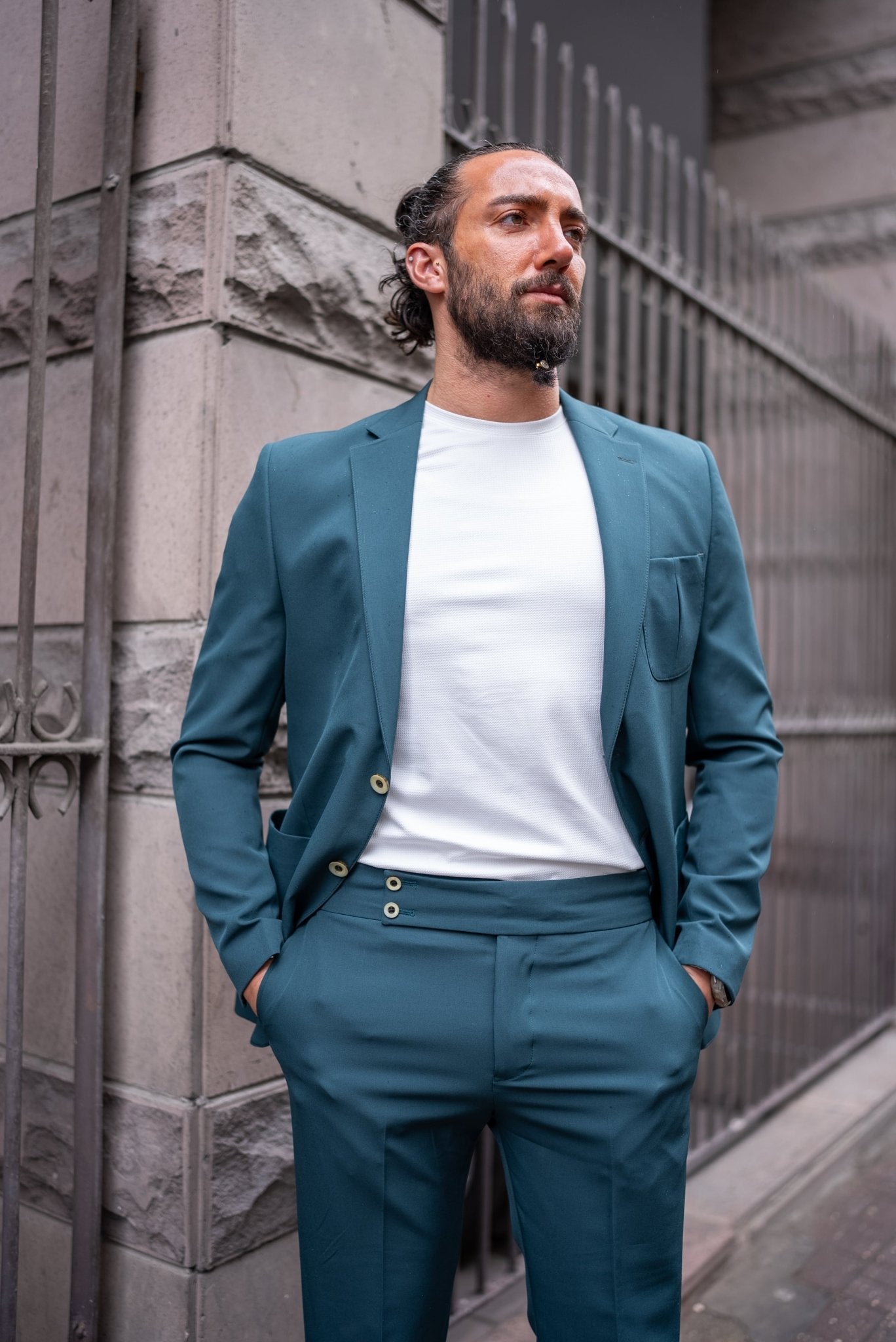 A man confidently models an Emerald Slim Fit Wedding-Casual Suit. The suit features a modern design with a sharp fit, highlighted by its vibrant emerald green color. He pairs the suit with a simple white shirt underneath, exuding a blend of elegance and casual sophistication. The man is accessorized with a large black ring, a watch, and minimal jewelry, standing in front of a stone building, capturing the essence of contemporary style.