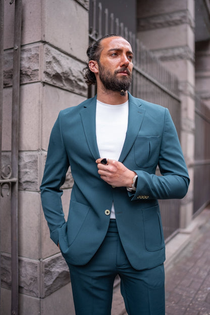 A man confidently models an Emerald Slim Fit Wedding-Casual Suit. The suit features a modern design with a sharp fit, highlighted by its vibrant emerald green color. He pairs the suit with a simple white shirt underneath, exuding a blend of elegance and casual sophistication. The man is accessorized with a large black ring, a watch, and minimal jewelry, standing in front of a stone building, capturing the essence of contemporary style.