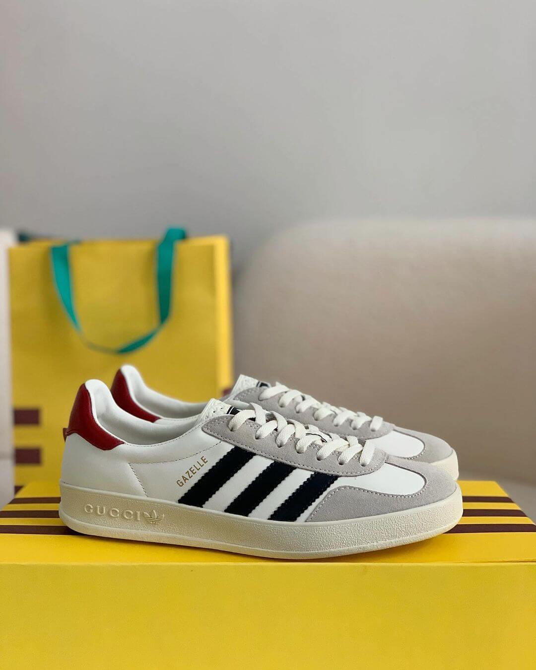 A pair of white Adidas x Gucci Gazelle sneakers with black stripes and a red heel tab, sitting on a yellow box with the Gucci logo.