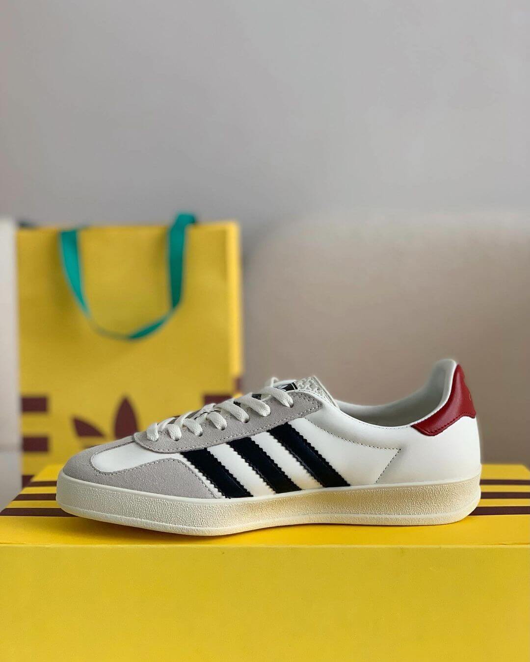 A pair of white Adidas x Gucci Gazelle sneakers with black stripes and a red heel tab