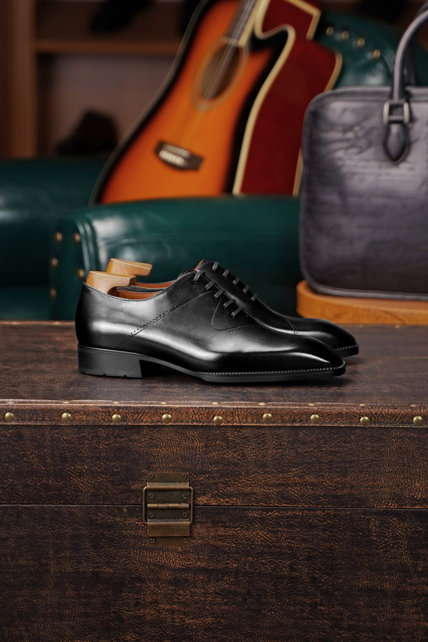 A pair of polished black leather oxford dress shoes with lace detailing, displayed on a dark wooden trunk in a styled setting with a green leather chair, an acoustic guitar, and a gray handbag in the background.