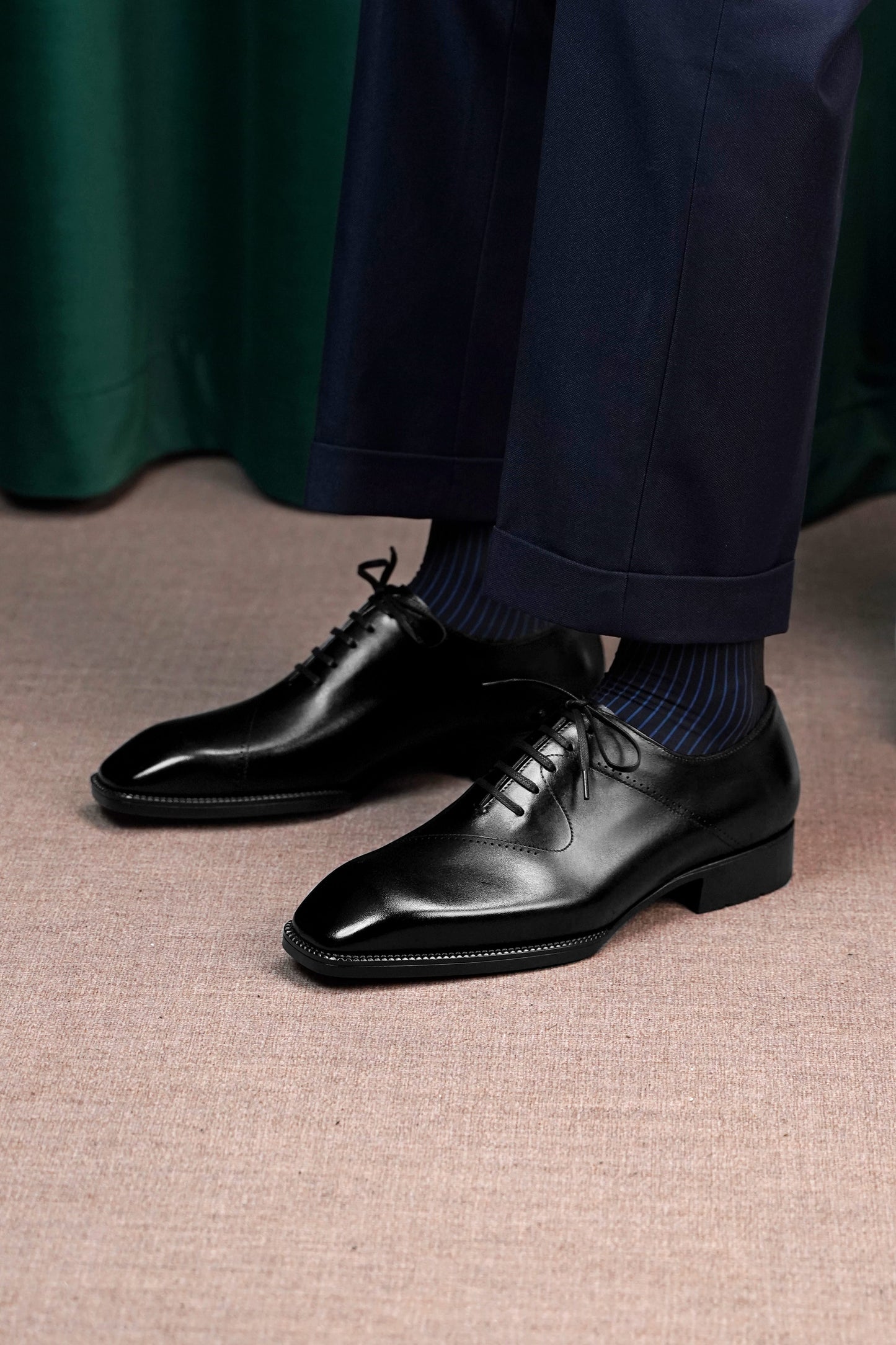 A pair of men's black leather oxford dress shoes with a cap toe, worn with dark blue ribbed socks and navy trousers, shown from a low angle on a textured beige floor.