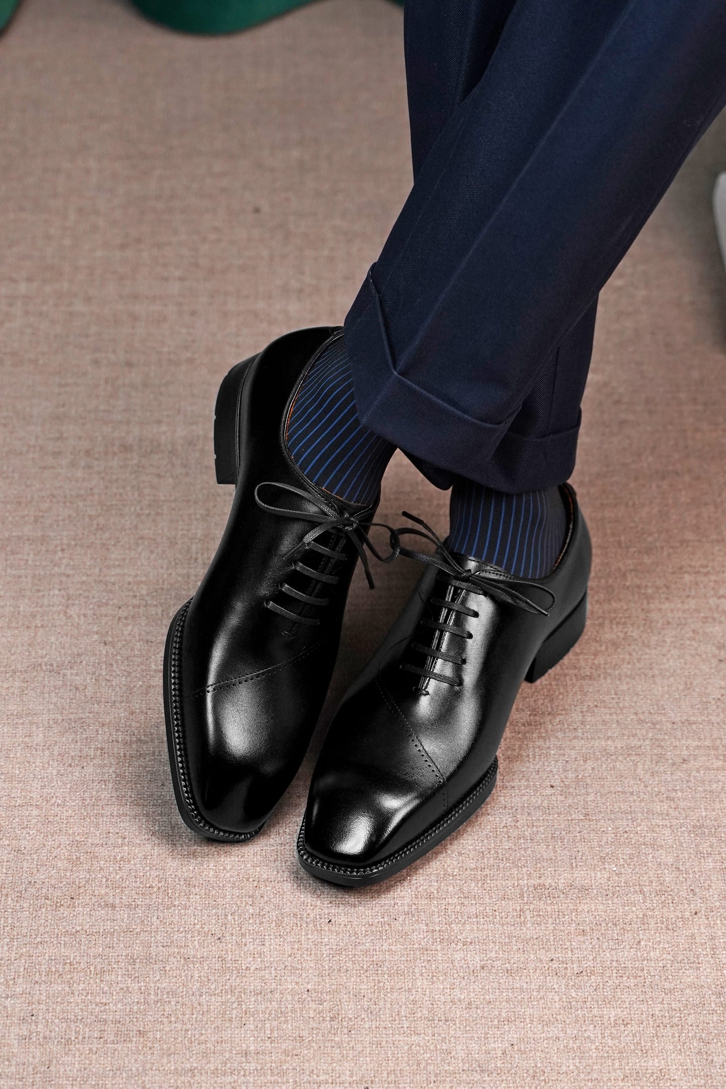 A pair of men's black leather oxford dress shoes with a cap toe, worn with dark blue striped socks and navy trousers on a textured beige carpet.