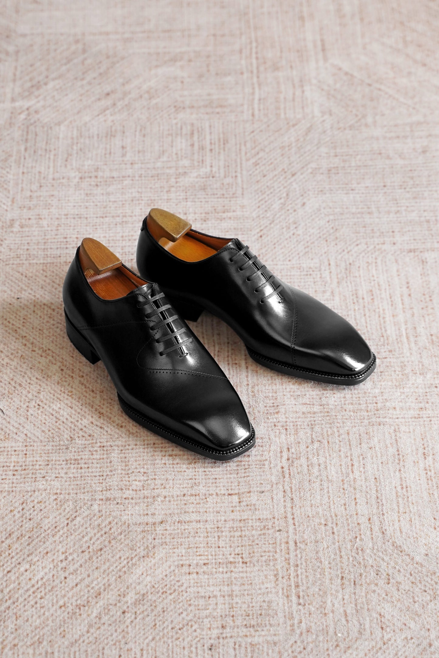 A pair of men's black leather oxford dress shoes with a wingtip toe and brogue detailing, displayed on a textured beige carpet.