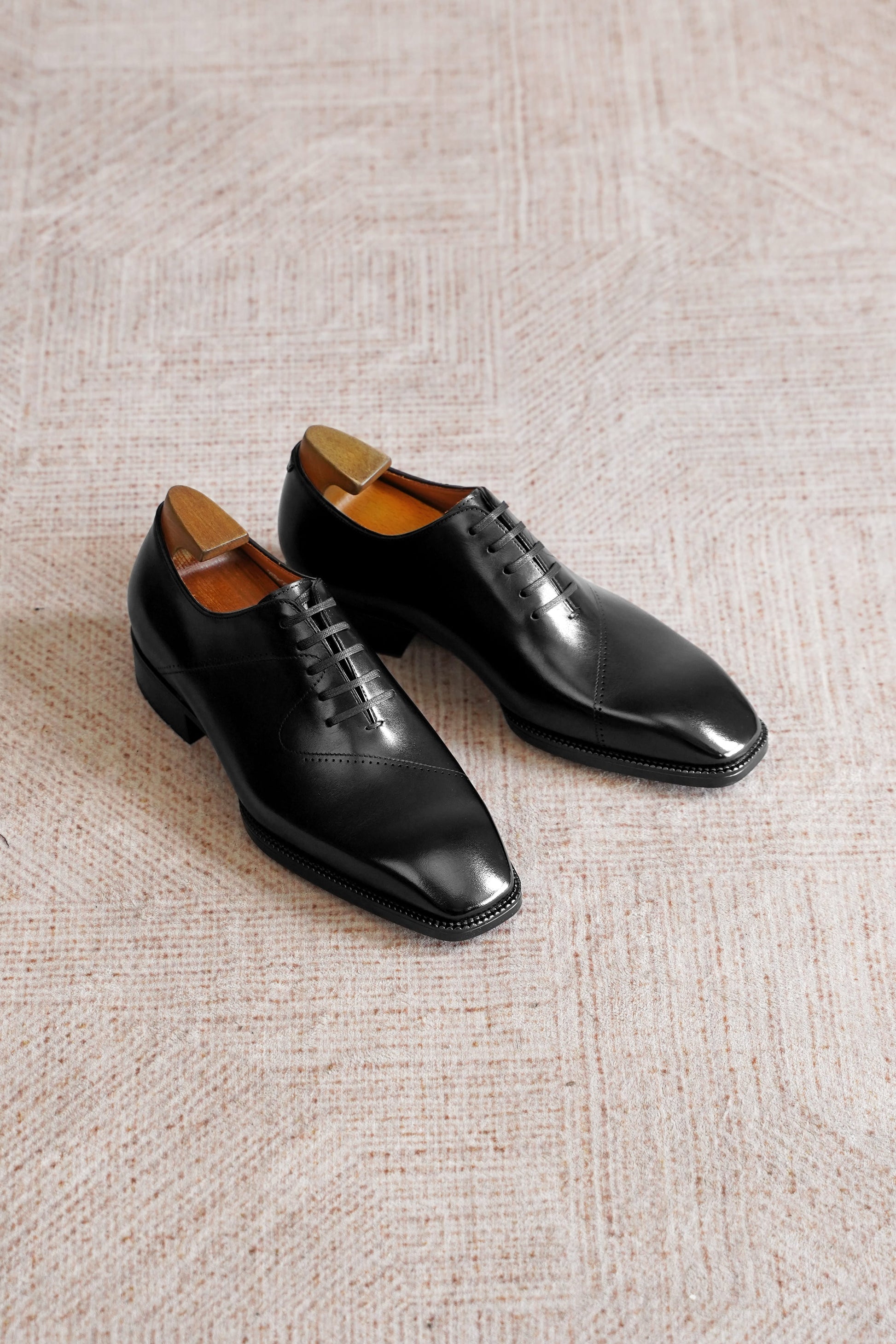 A pair of men's black leather oxford dress shoes with a wingtip toe and brogue detailing, displayed on a textured beige carpet.