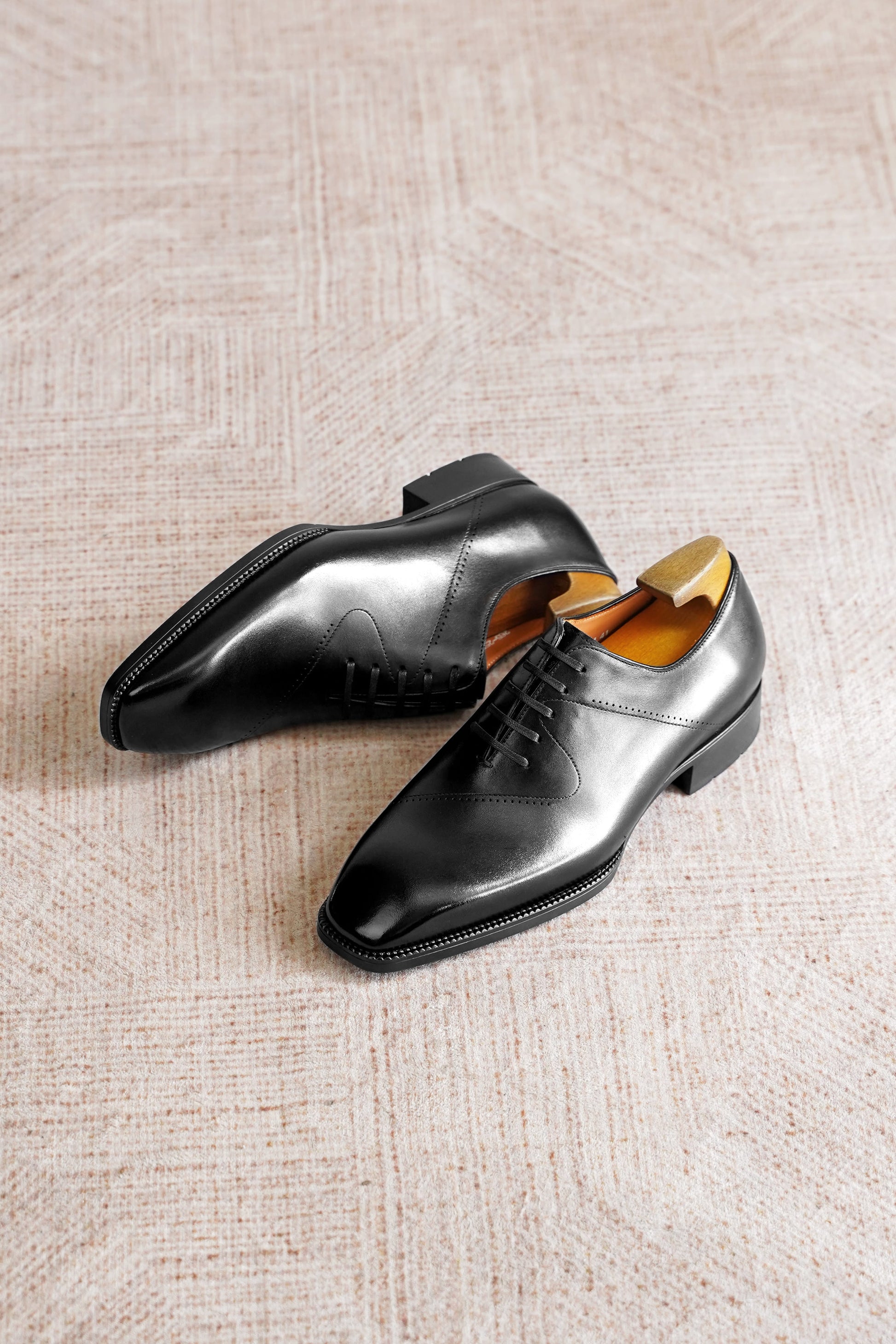 A pair of black leather dress shoes with laces, displayed on a textured beige woven rug. One shoe is positioned upright, showing its side profile and black sole, while the other lies on its side, revealing the tan interior lining and a wooden shoe tree inserted inside.