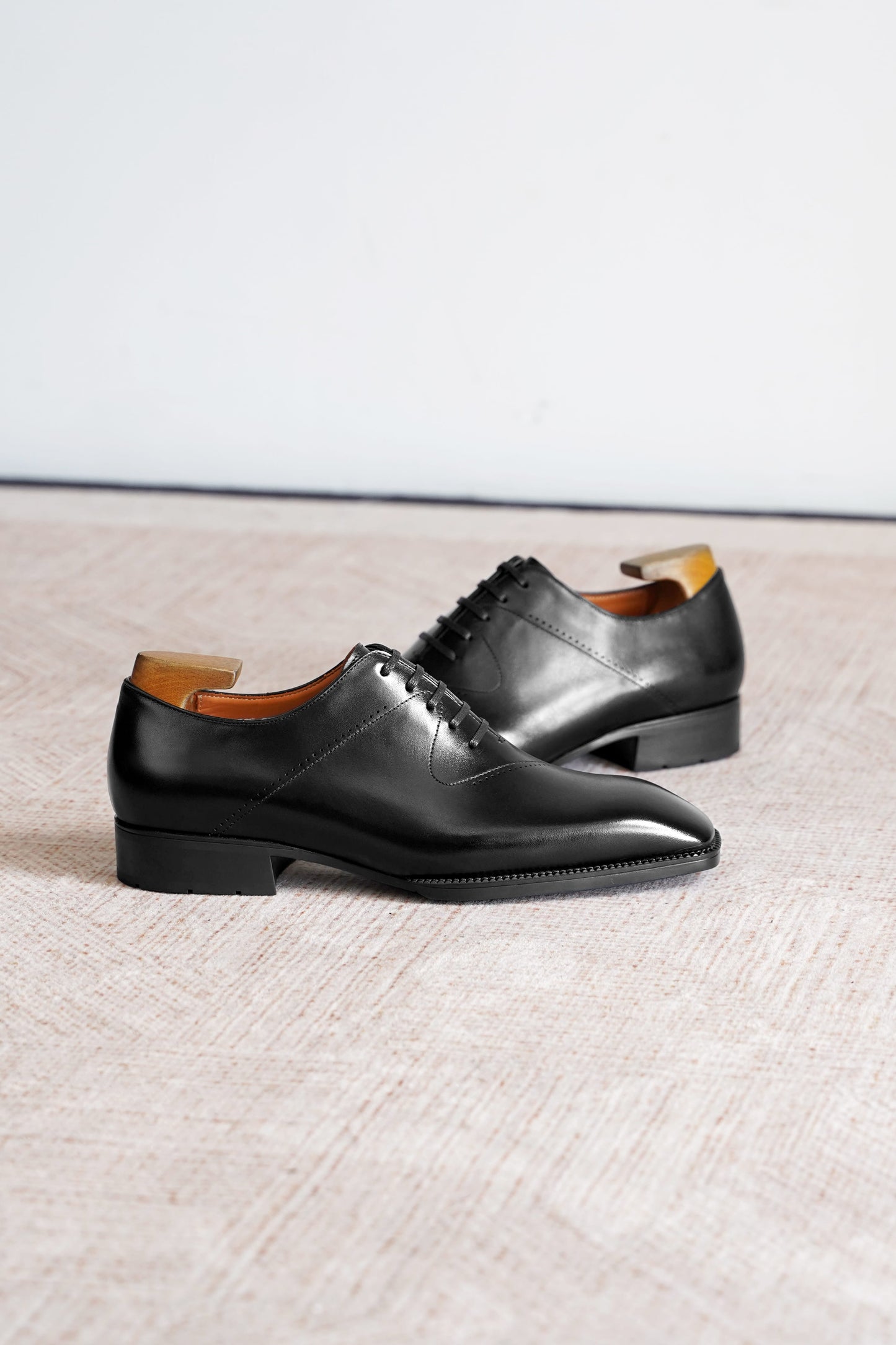 A pair of black leather oxford dress shoes with a low stacked heel and pointed toe, displayed on a textured beige surface against a light gray background.