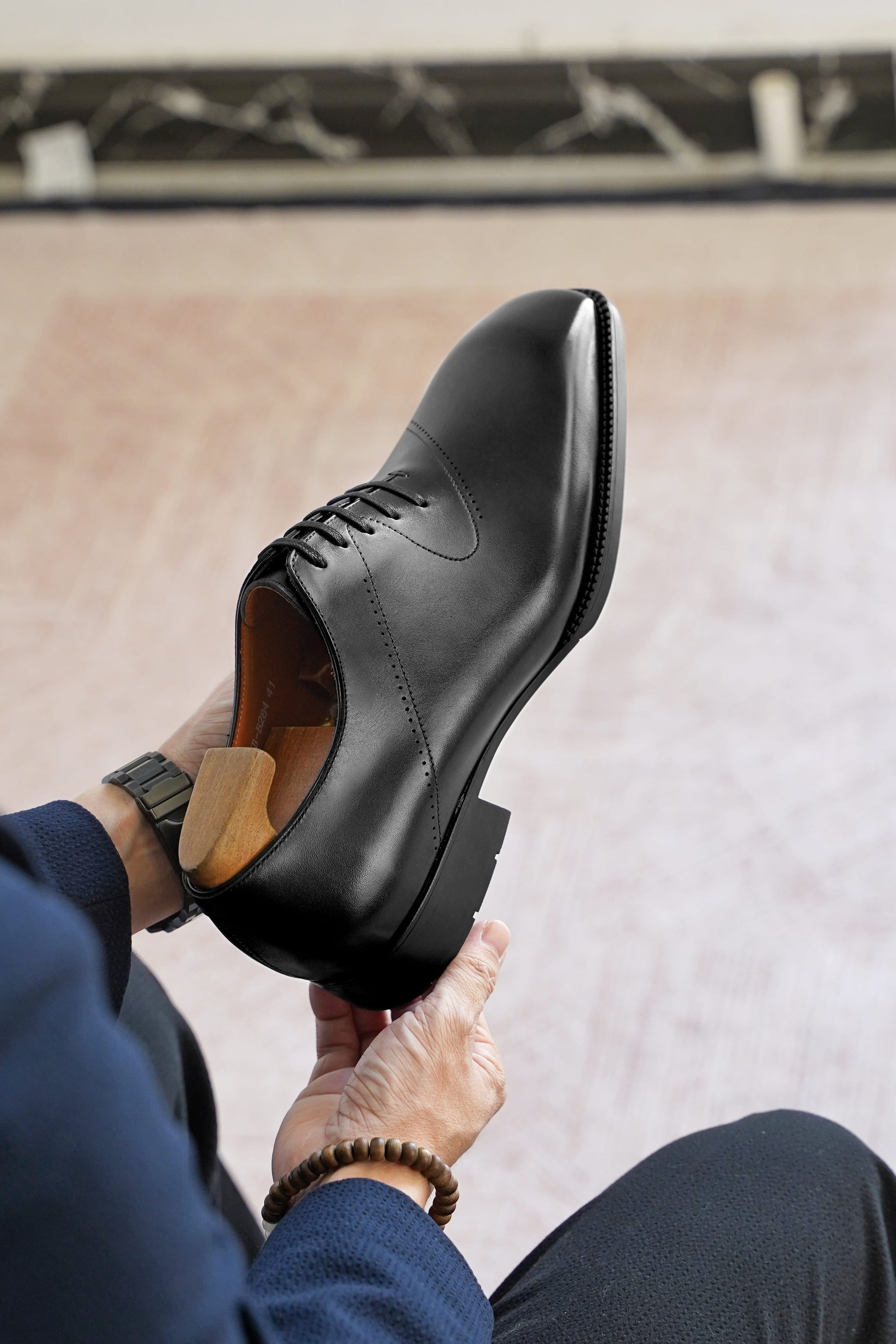 A person holds up a black leather dress shoe with a wingtip toe and brogue detailing, showing the side profile and interior lining. The person is wearing a navy blue sweater and dark trousers, with a wooden shoe tree inside the shoe.
